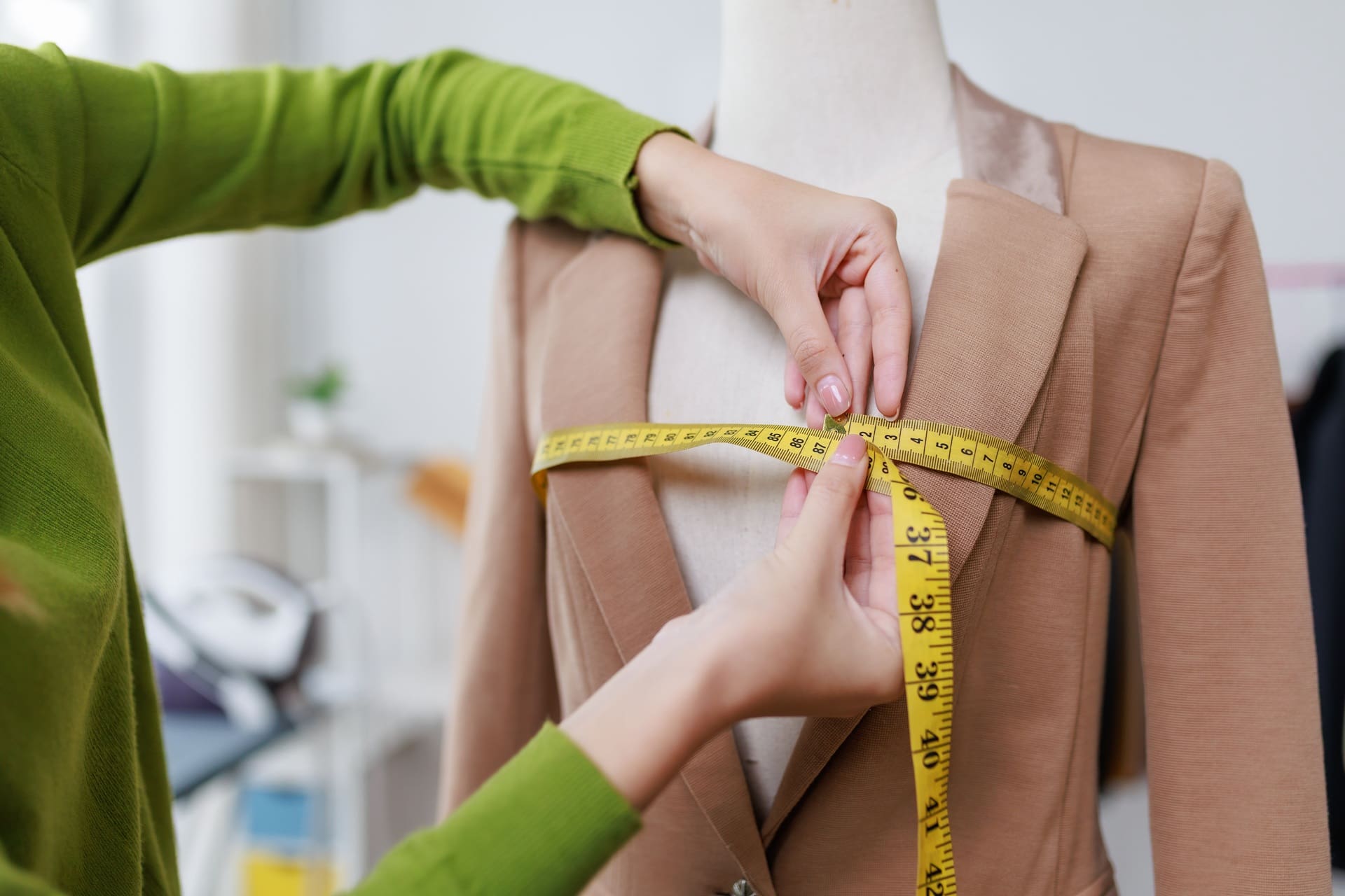 A person in a green sleeve measures the chest area of a beige blazer on a dress form using a yellow measuring tape.