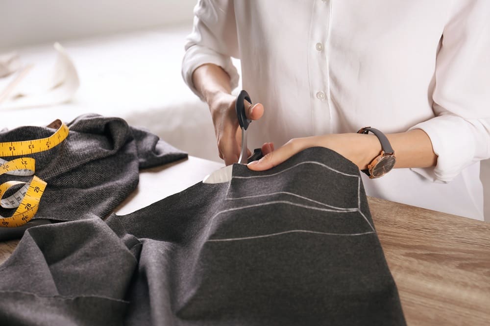 Person cutting dark fabric with scissors on a table, measuring tape nearby, and chalk lines visible on the cloth.