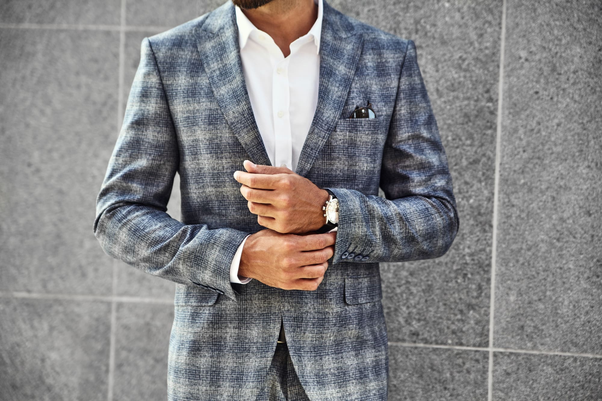 A man in a checked grey suit and white shirt adjusts his sleeve while standing against a textured grey wall.