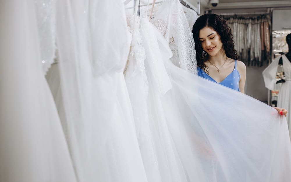 A woman in a blue dress examines a white wedding gown in a bridal shop, surrounded by other white dresses on hangers.